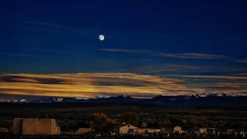 A moonrise over a small New Mexico village at dusk, with adobe buildings and a church in the foreground, autumn cottonwoods, a cemetery, and snow-capped Sangre de Cristo mountains along the horizon beneath a deep blue sky streaked with golden sunset clouds.
