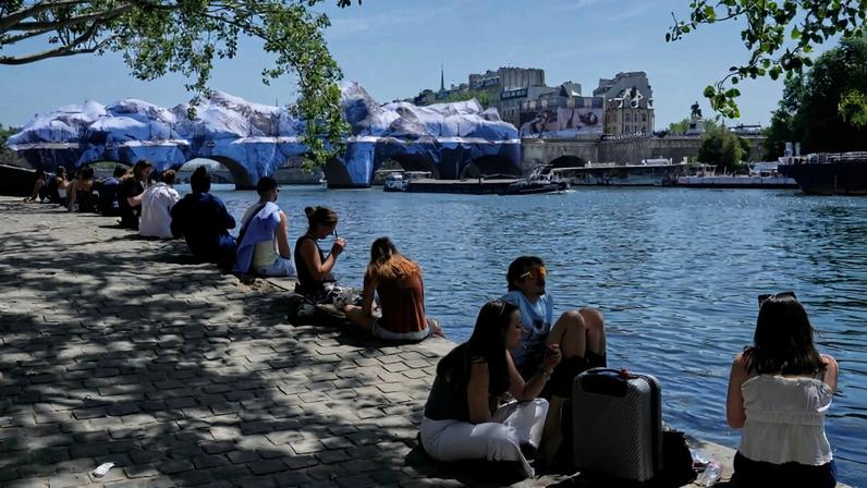People seek relief from the heat along the Seine River in Paris, Monday, May 25, 2026. Artwork by street artist JR is seen on the Pont Neuf in the background.