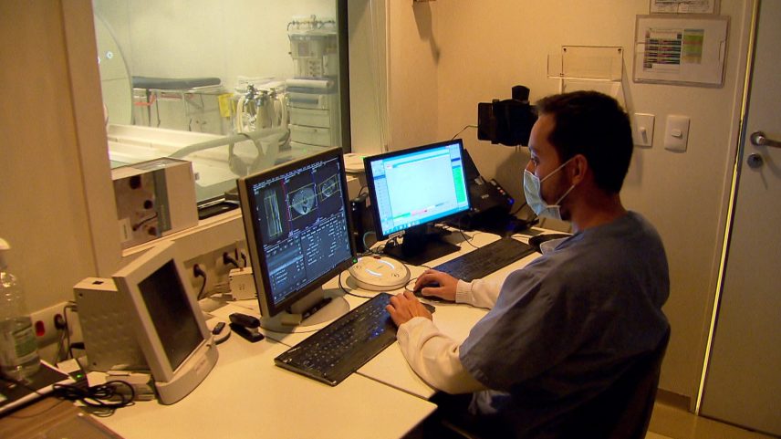 Profissional de saúde utiliza computador em sala de controle enquanto monitora exames em equipamento hospitalar