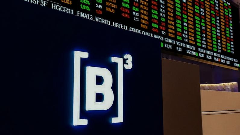 Logo da B3 em tela azul, com painel de cotações da bolsa de valores exibindo dados financeiros ao fundo.