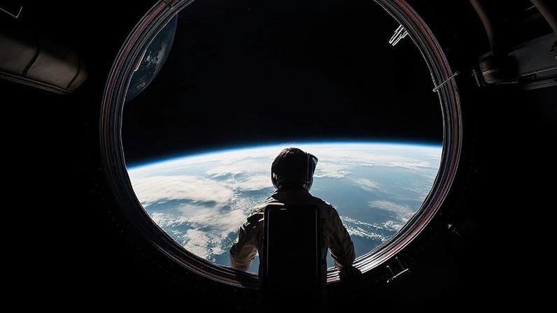 Pessoa de costas observa a Terra com nuvens e o espaço através de uma escotilha circular de nave espacial.
