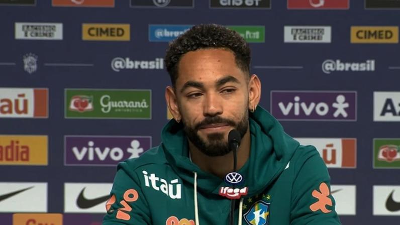 Homem negro de barba e cabelo crespo, vestindo moletom verde da seleção brasileira, em coletiva de imprensa com fundo de patrocinadores