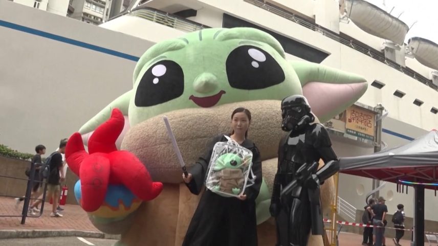 A imagem mostra uma cena de evento temático ao ar livre, provavelmente uma convenção geek ou de cultura pop. No centro, uma pessoa segura um sabre de luz e uma bolsa transparente com um boneco do Grogu (Baby Yoda), personagem de Star Wars.