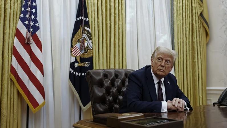O presidente dos EUA, Donald Trump, em terno escuro, está sentado no Salão Oval, com bandeiras dos EUA e do Exército ao fundo, adornado com medalhas e uma estátua de cavalo na mesa.