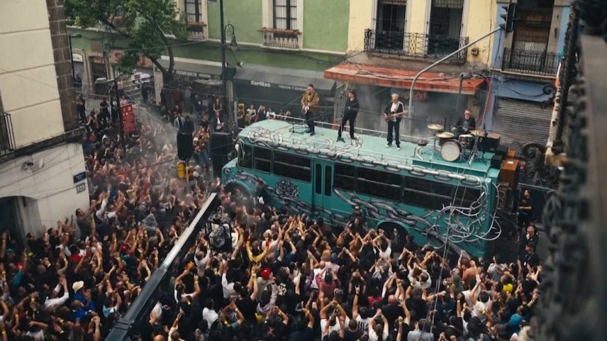 Banda em cima de ônibus turquesa cercado por grande multidão em rua urbana.