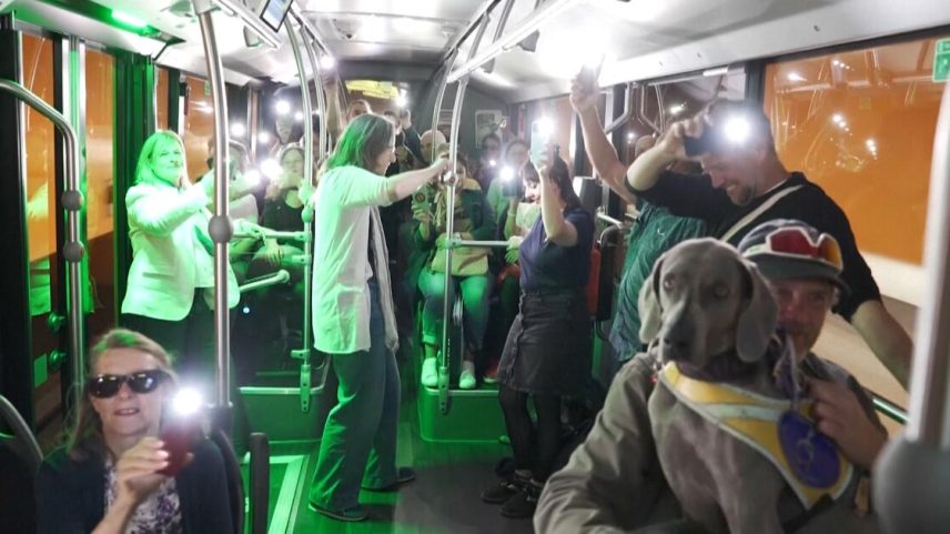 Interior de ônibus iluminado com luz verde, cheio de pessoas segurando celulares com lanterna acesa; clima festivo e descontraído.