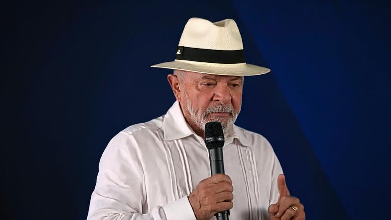 O presidente Lula tem um dedo apontado para cima e usa umm chápeu e uma camisa branca
