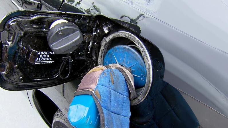 Um frentista branco insere uma mangueira azul de gasolina no bocal de abastecimento de um carro preto. O reflexo do trabalhador pode ser observado na pintura do automóvel,