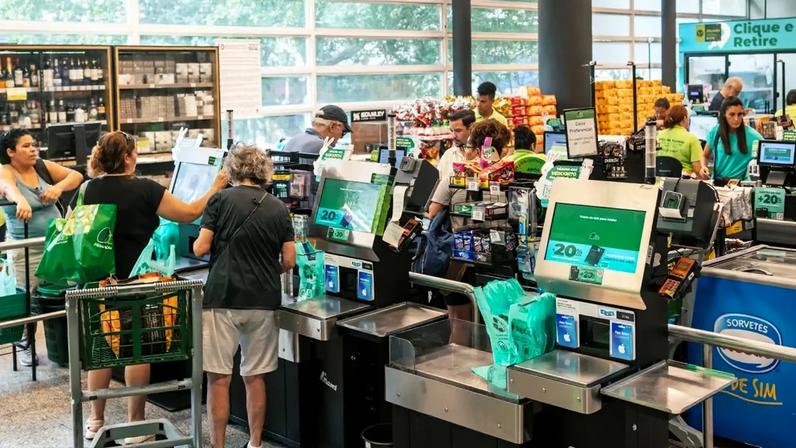 Interior de supermercado com caixas de autoatendimento e tradicionais, pessoas usando carrinhos de compras ou sacolas verdes, iluminação natural e prateleiras com lanches e bebidas; funcionários uniformizados em camisetas verdes.