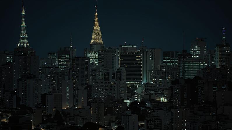 Vista noturna da cidade de São Paulo, com dois edifícios em formato de torre iluminados no topo, destacando-se entre os demais prédios residenciais e comerciais com janelas acesas, sob um céu escuro