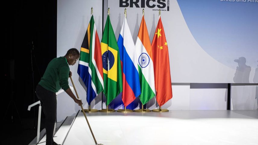 Imagem colorida mostra homem varrendo o chão, com bandeiras dos países do Brics ao fundo - Metrópoles