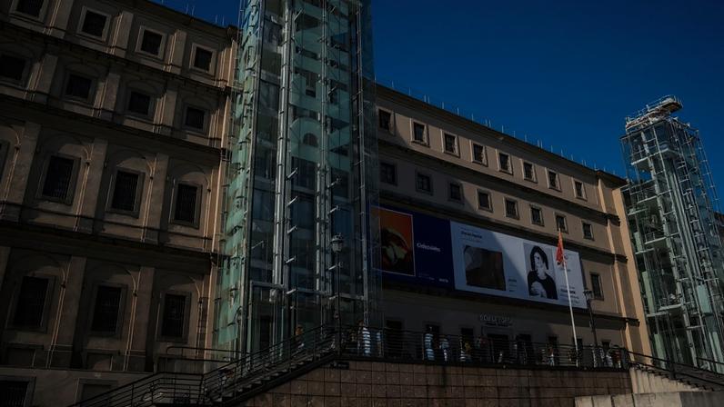 Visitors line the stairs leading to the Reina Sofía museum in Madrid, a long official looking four story building, set against a blue sky.