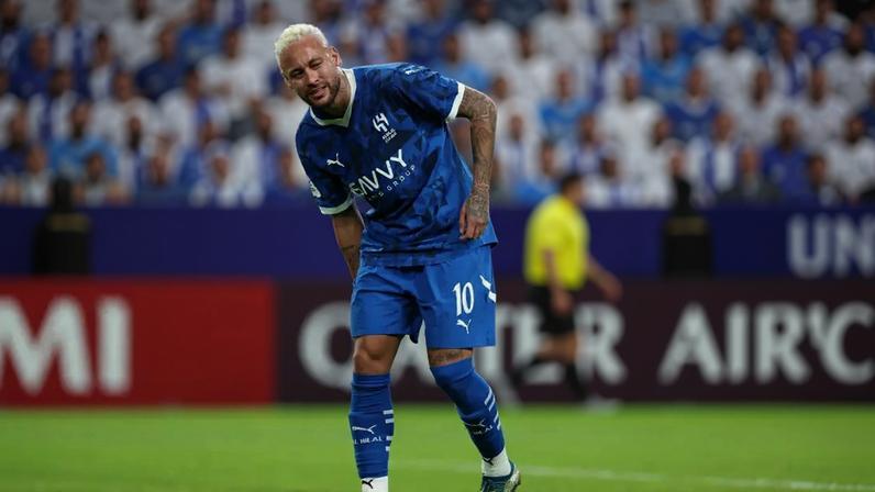 Neymar Jr. em campo, vestindo uniforme azul com o número 10, cabelo loiro platinado, expressão séria, inclinando-se levemente para frente, com as mãos nos joelhos, em um estádio de futebol com torcedores ao fundo