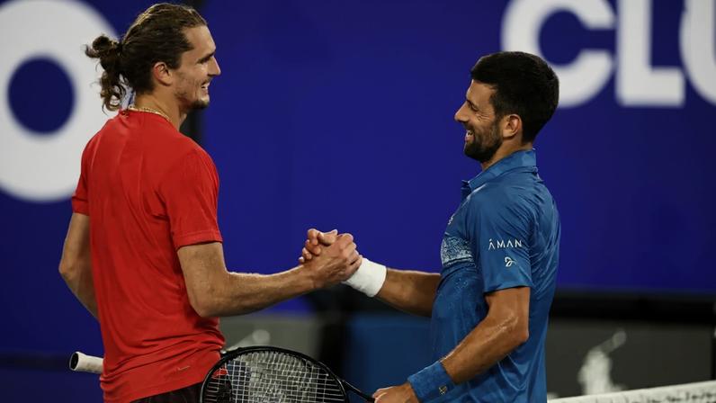 Alexander Zverev and Novak Djokovic shake hands over a tennis net holding their rackets.