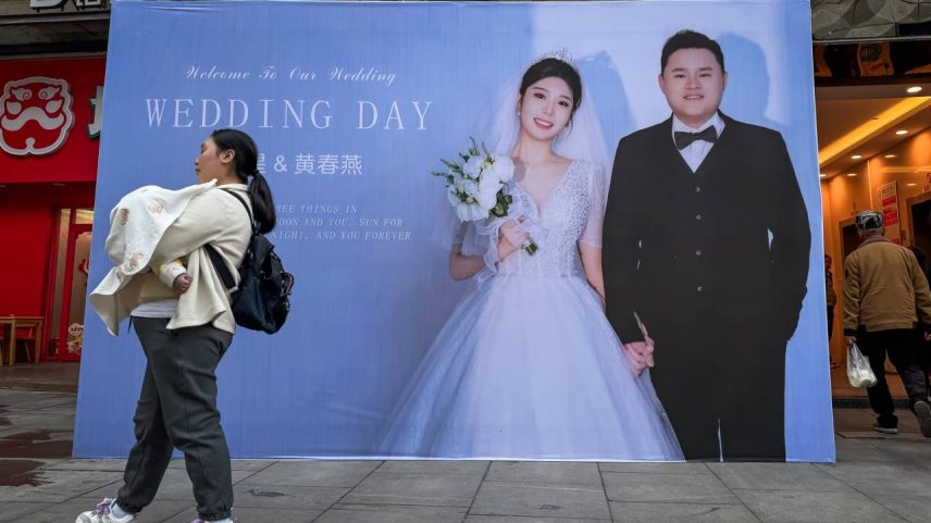 Uma mulher carregando uma criança passa por um grande anúncio de casamento na entrada de um hotel, indicando que o casal retratado no outdoor provavelmente realizará seu banquete de casamento ali. À medida que as taxas de casamento e natalidade na China continuam a diminuir, cenas como essa destacam o contraste entre a cultura tradicional de casamento e as mudanças demográficas e tendências sociais em 10 de março de 2025 em Chongqing, China