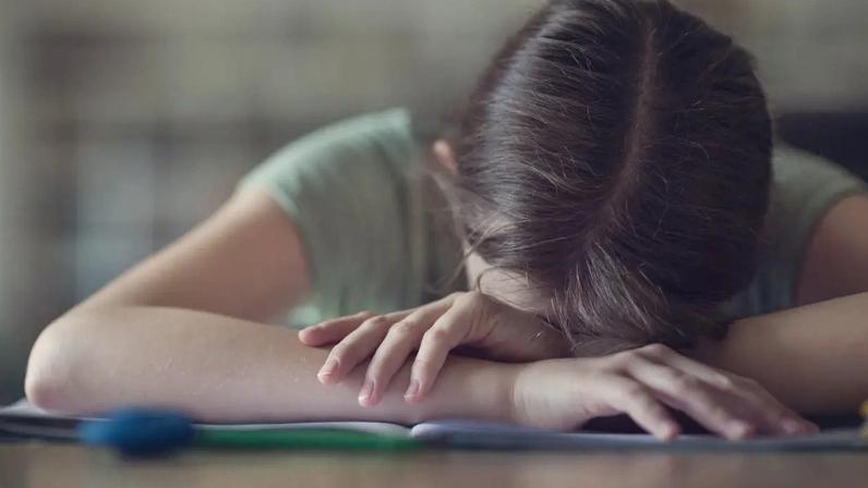 Adolescente com cabelo castanho e camiseta verde deitada sobre os braços em uma mesa, com um caderno aberto e canetas coloridas à frente, transmitindo cansaço ou desânimo