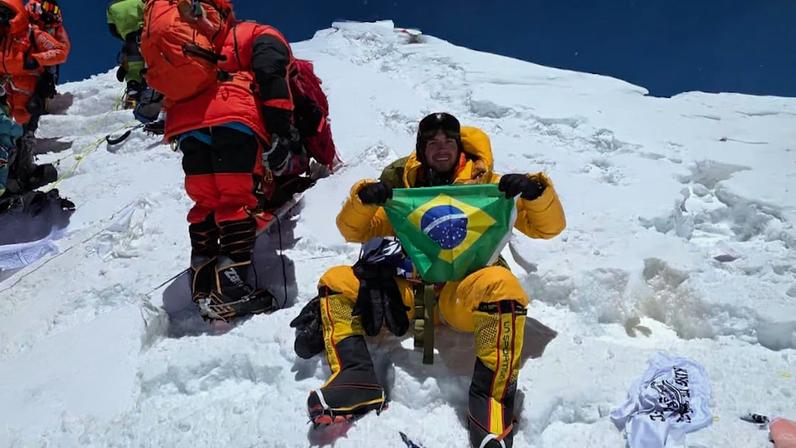Um jovem de 24 anos sorri no topo do Everest e segura a bandeira do Brasil.