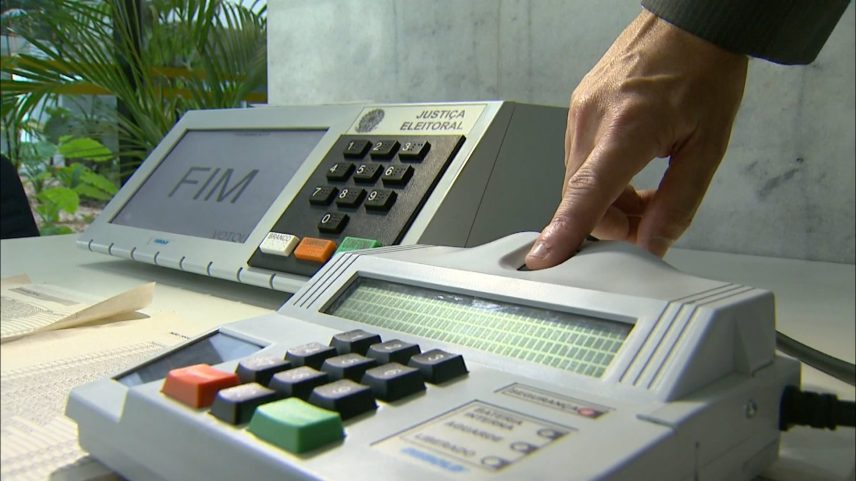 Urna eletrônica brasileira com tela exibindo “FIM” e mão pressionando botão; indica encerramento da votação.