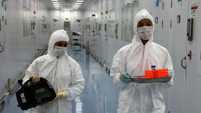 Duas pessoas com traje de biossegurança com capuz e máscaras médicas no rosto segurando equipamentos laboratoriais num corredor