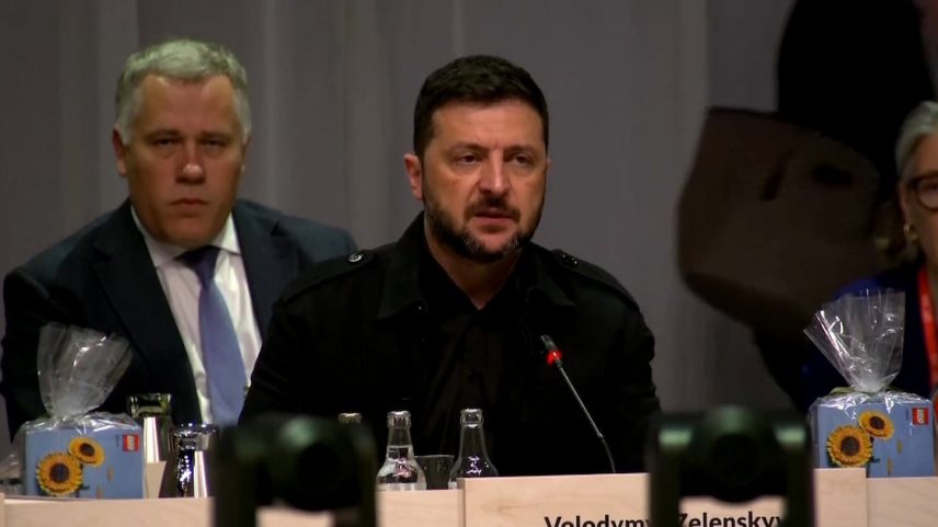 Volodymyr Zelensky em conferência, falando ao microfone, sentado à mesa com garrafas e participantes ao fundo