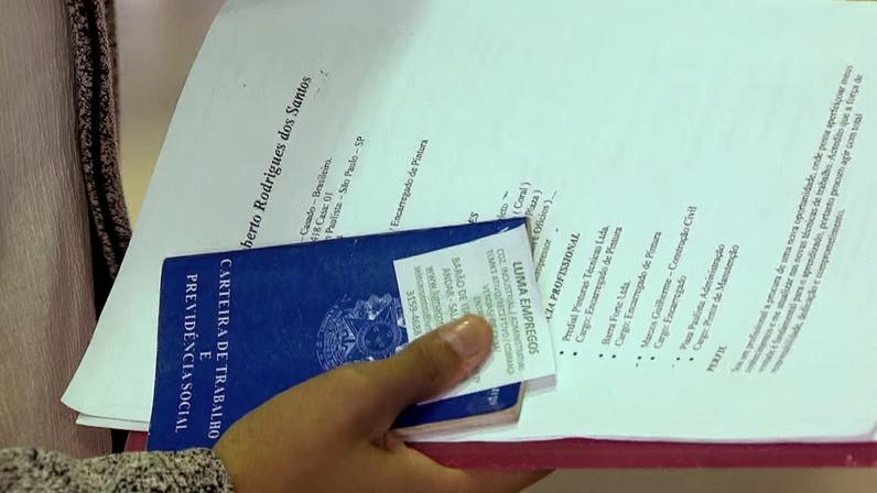 Pessoa segura documentos de candidatura a emprego. Entre eles, há uma Carteira de Trabalho e Previdência Social azul, um currículo e um cartão de agência de empregos. Os papéis estão presos em uma pasta vermelha.