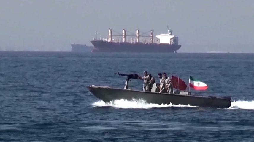 Embarcação militar com soldados armados no mar, com navios cargueiros ao fundo