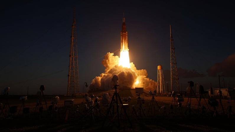 NASA's SLS (Space Launch System) rocket rises over the launch pad and into the blue sky, with fire and smoke underneath it. In front of it are 20 to 30 cameras to capture the historic moment.