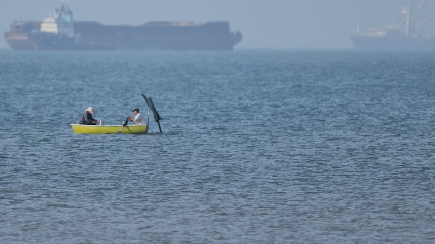 Dois homens estão sentados em um pequeno barco na água, enquanto navios cargueiros estão ancorados ao fundo no Estreito de Ormuz