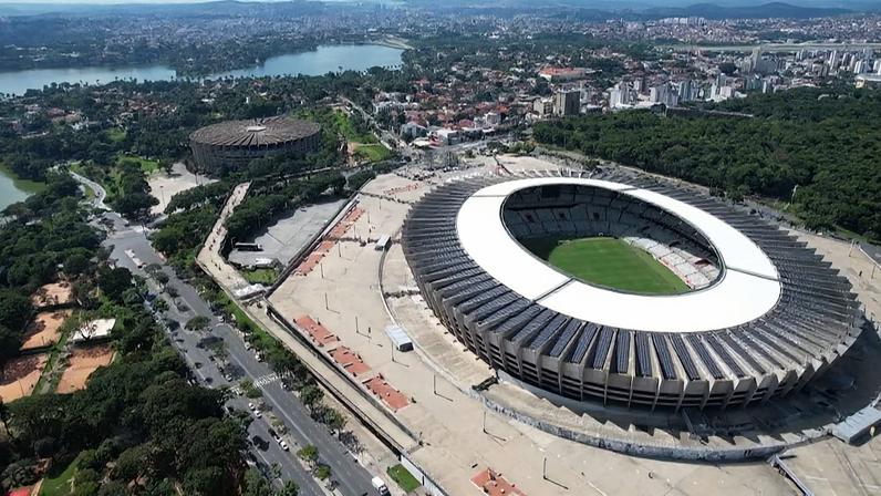 Vista aérea de grande estádio circular com campo verde e cobertura branca cercada por painéis solares.