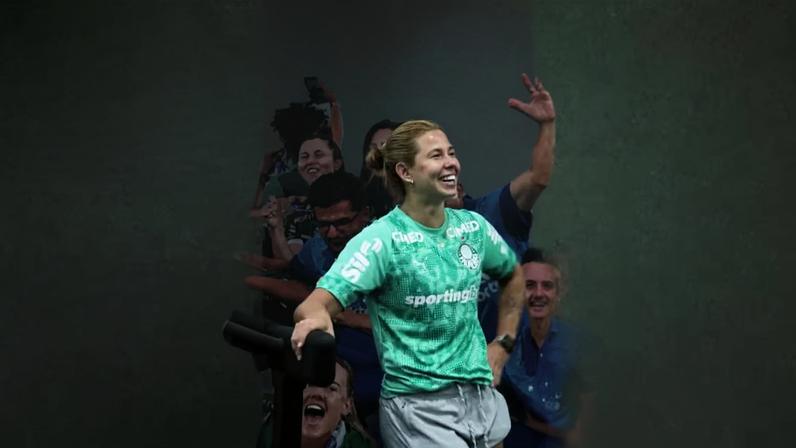 Atleta com uniforme verde e shorts cinza sorrindo diante de mural com torcedores ao fundo.