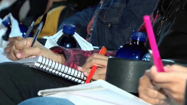 Pessoa escrevendo em caderno com caneta durante aula, ao lado de garrafas de água e outros estudantes fazendo anotações
