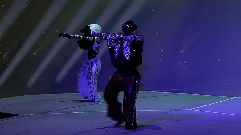 Dois robôs em palco iluminado por luzes coloridas, vestindo figurinos brilhantes.