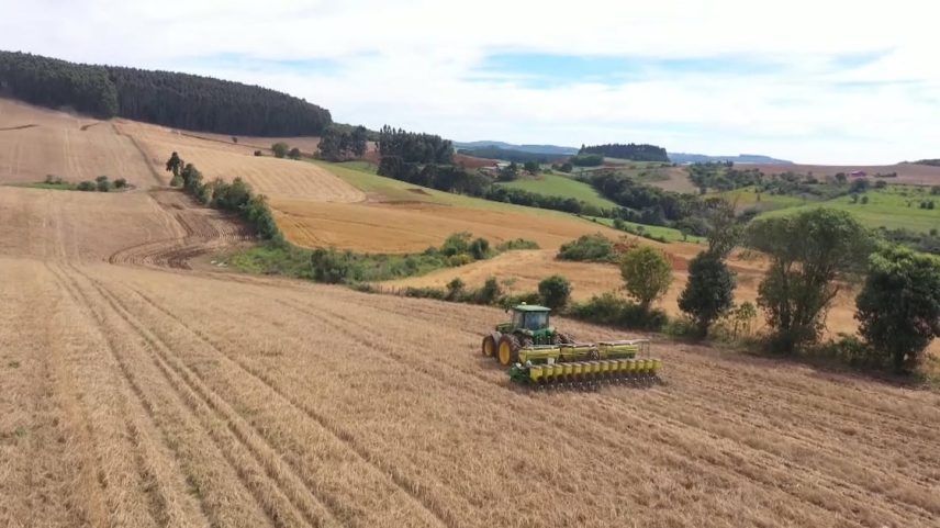 Trator verde trabalha em campo de colheita cercado por morros e vegetação rural.
