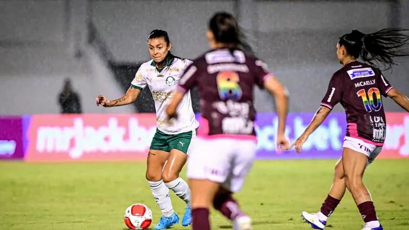 Jogo de futebol feminino em campo gramado. A jogadora do time de uniforme branco e verde domina a bola vermelha e branca, enquanto duas adversárias com uniforme vinho e se aproximam para disputar a jogada.