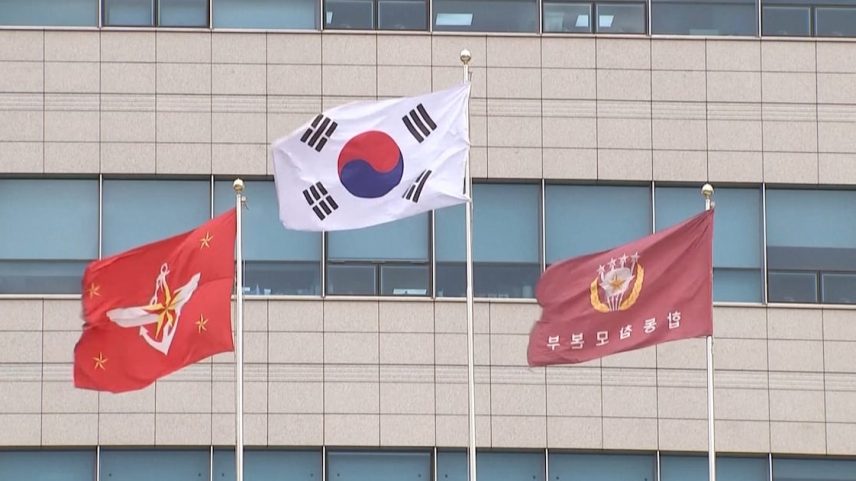 A bandeira da Coreia do Sul está içada no mais alto dos mastros em frente a um prédio. Ao lado dela, encontram-se bandeiras militares,