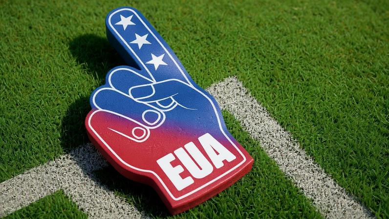 Ilustração 3D de uma mão de espuma de torcida com as cores da bandeira dos EUA e a sigla "EUA" em branco, sobre gramado verde na marca de escanteio.