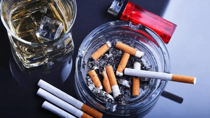 Image, vista de cima, de um cinzeiro com várias bitucas de cigarros e um copo de whisky com pedras de gelo. Ao lado vê-se um isqueiro vermelho e três cigarros intocados.