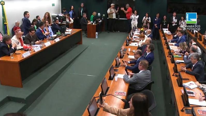Reunião formal na Câmara dos Deputados com várias pessoas sentadas em mesas longas, usando computadores e discutindo durante uma sessão