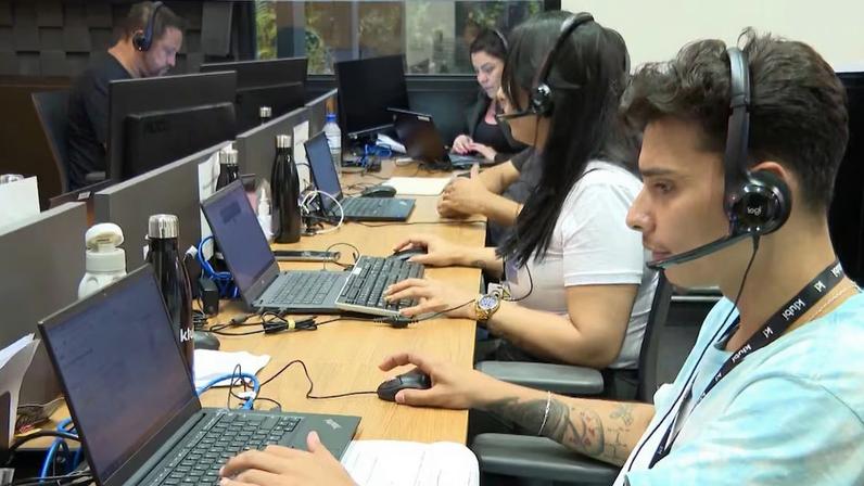 Em uma longa mesa de madeira, diversas garrafas e notebooks estão posicionados. Um jovem com tatuagens no braço direito e um fone de ouvido trabalha em um dos laptops. Ao lado dele, colegas fazem o mesmo.