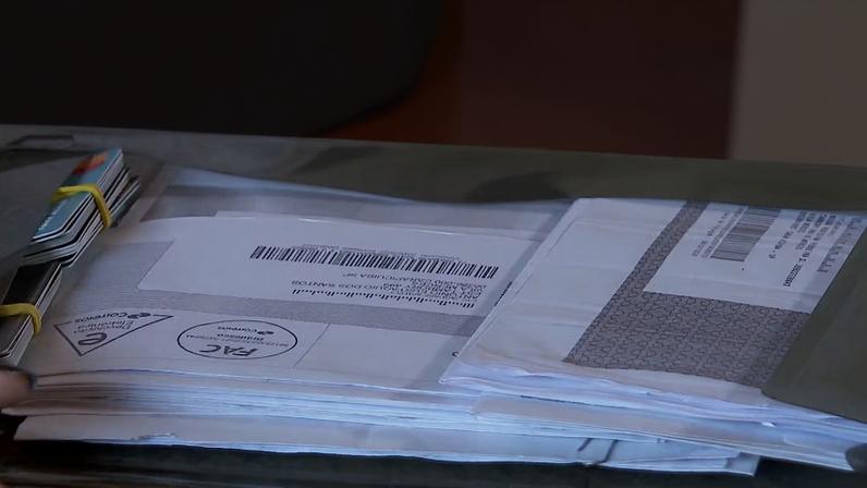 Pilha de documentos e envelopes com códigos de barras sobre a mesa, sendo organizada por uma mão