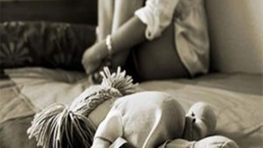 Foto mostra criança retraída no canto com boneca na frente.