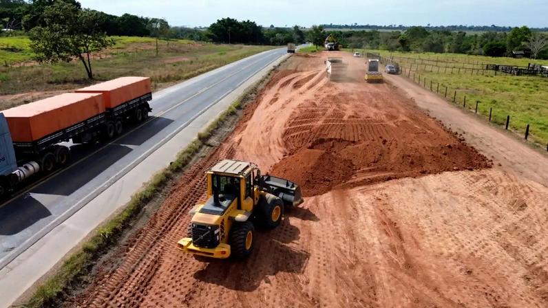 Vista aérea de uma rodovia com um caminhão azul e laranja à esquerda, e à direita, uma área em construção com terra vermelha, onde uma pá carregadeira amarela move terra e outros veículos trabalham ao fundo