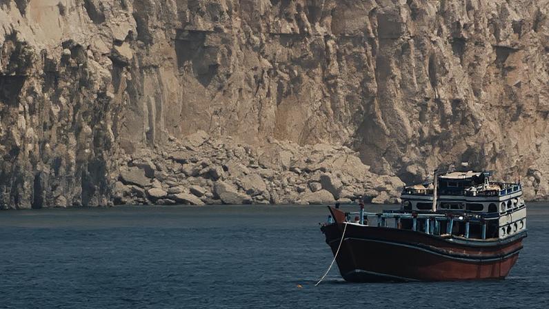 Um barco está ancorado no meio do Estreito de Ormuz. Atrás dele é possível ver um grande paredão de pedra.