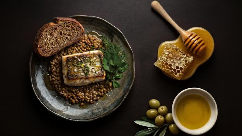 Prato com peixe grelhado sobre lentilhas, pão integral e ervas frescas. Ao lado, mel com favo, azeite em tigela e azeitonas verdes com folhas