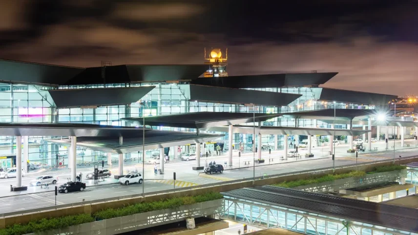 Entrada do Aeroporto de Guarulhos, com estruturas de vidro, torre de controle iluminada, telhado dinâmico, via movimentada e iluminação intensa.