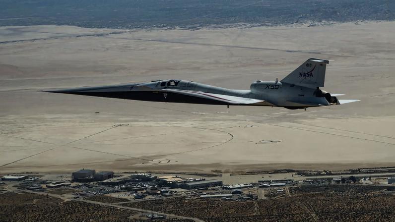 NASA’s X-59 quiet supersonic research aircraft flies above NASA’s Armstrong Flight Research Center in Edwards, California. The plane has a distinctive shape, with a very long, sharp nose.