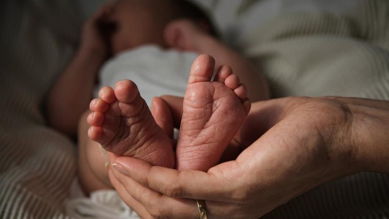 1 de 1 Imagem colorida de um bebê vestindo fraldas brancas e deitado sobre um lençol branco. Uma mão com uma aliança dourada segura os pés dele - Foto: Rene Terp/Pexels