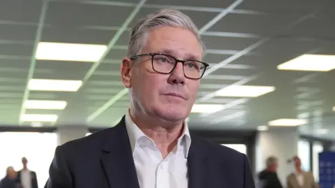Sir Keir Starmer, in a white open necked shirt and dark suit jacket, wearing glasses and making a serious expression. He appears inside a venue with the faces of people in the background blurred