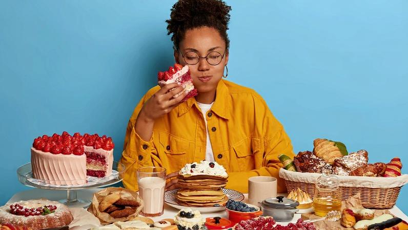 Foto colorida de mulher sentada atrás de uma mesa com muitas comidas, segurando um pedaço enorme de bolo com as mãos -Metrópoles.