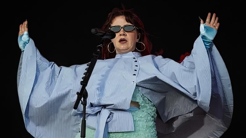 CMAT performs on Radio 1's Big Weekend stage against a pink backdrop with yellow stars. She wears a light blue floral two-piece outfit with a blue striped cape and has shoulder length red hair. She wears silver hoops and sunglasses and holds up her hands to the crowd